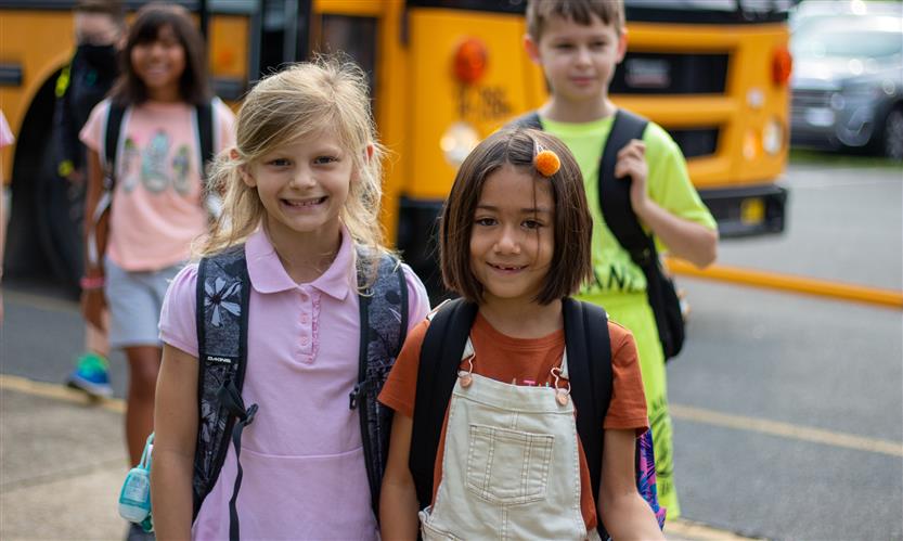 Two students smile while standing in front of a school bus